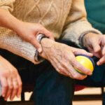 An elderly person receives support from a caregiver, holding hands indoors, showcasing compassion.