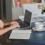 Female freelancer using laptop with coffee at home office desk, surrounded by roses and a planner.