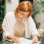 Woman analyzing financial documents using laptop and calculator indoors.