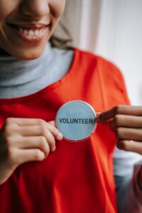 Crop anonymous positive ethnic female smiling and demonstrating sign with inscription volunteer on bright red apron